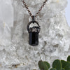 Light reflects off the faces of a black cylindrical Tourmaline stone in an antiqued copper setting, he pendant hangs from a copper chain. Green eucalyptus leaves are arranged below a Quartz crystal cluster.