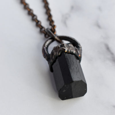 Light reflects off the face of a black cylindrical Tourmaline stone in an antiqued copper setting, the pendant hangs from a copper chain.