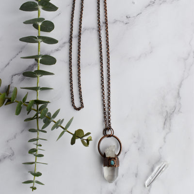 A Clear Quartz point crystal is encircled with antique copper and accented with a small hexagonal blue flash Labradorite stone. The necklace is displayed beside green eucalyptus leaves and a Quartz point are arranged nearby.