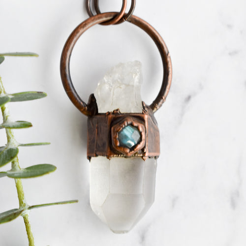 A Clear Quartz point crystal is encircled with antique copper and set with a small hexagonal blue flash Labradorite stone. Green eucalyptus leaves are arranged nearby.