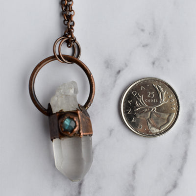 A Clear Quartz point crystal is encircled with antique copper and accented with a small hexagonal blue flash Labradorite stone, and hangs from a rolo chain. The necklace is displayed beside a Quarter coin for size reference.