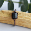 A Black Tourmaline and copper ring is displayed over a stick of Palo Santo wood. Eucalyptus leaves are in the background.