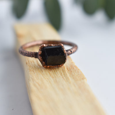 A Black Tourmaline and copper ring is displayed over a stick of Palo Santo wood. Eucalyptus leaves are in the background.