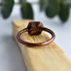 The textured and antiqued copper back of a ring is shown displayed on top of a Palo Santo sick of wood. Green eucalyptus leaves fade into the background.