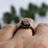 Black stone and copper ring is worn on hand. Green leaves in background.