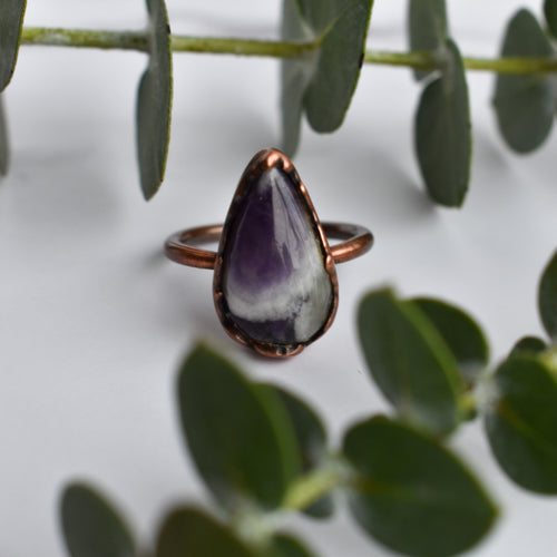 Purple, white and grey Chevron Amethyst stone in an antiqued copper ring, shown between eucalyptus leaves.