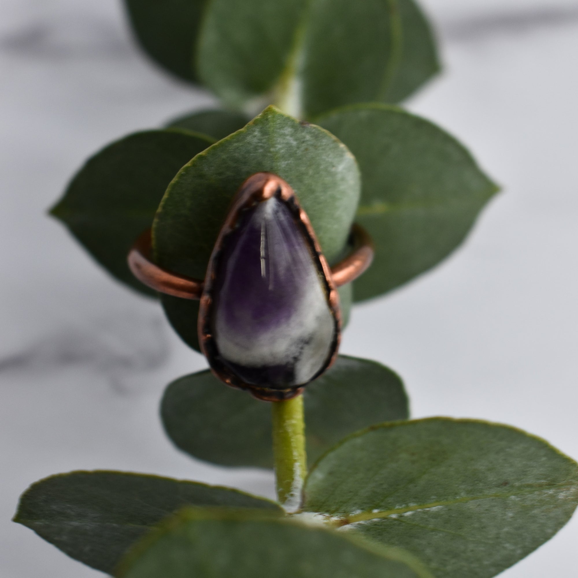 Tear dropped shaped purple, white and grey crystal ring. Displayed over a eucalyptus leaf.