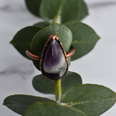 Tear dropped shaped purple, white and grey crystal ring. Displayed over a eucalyptus leaf.