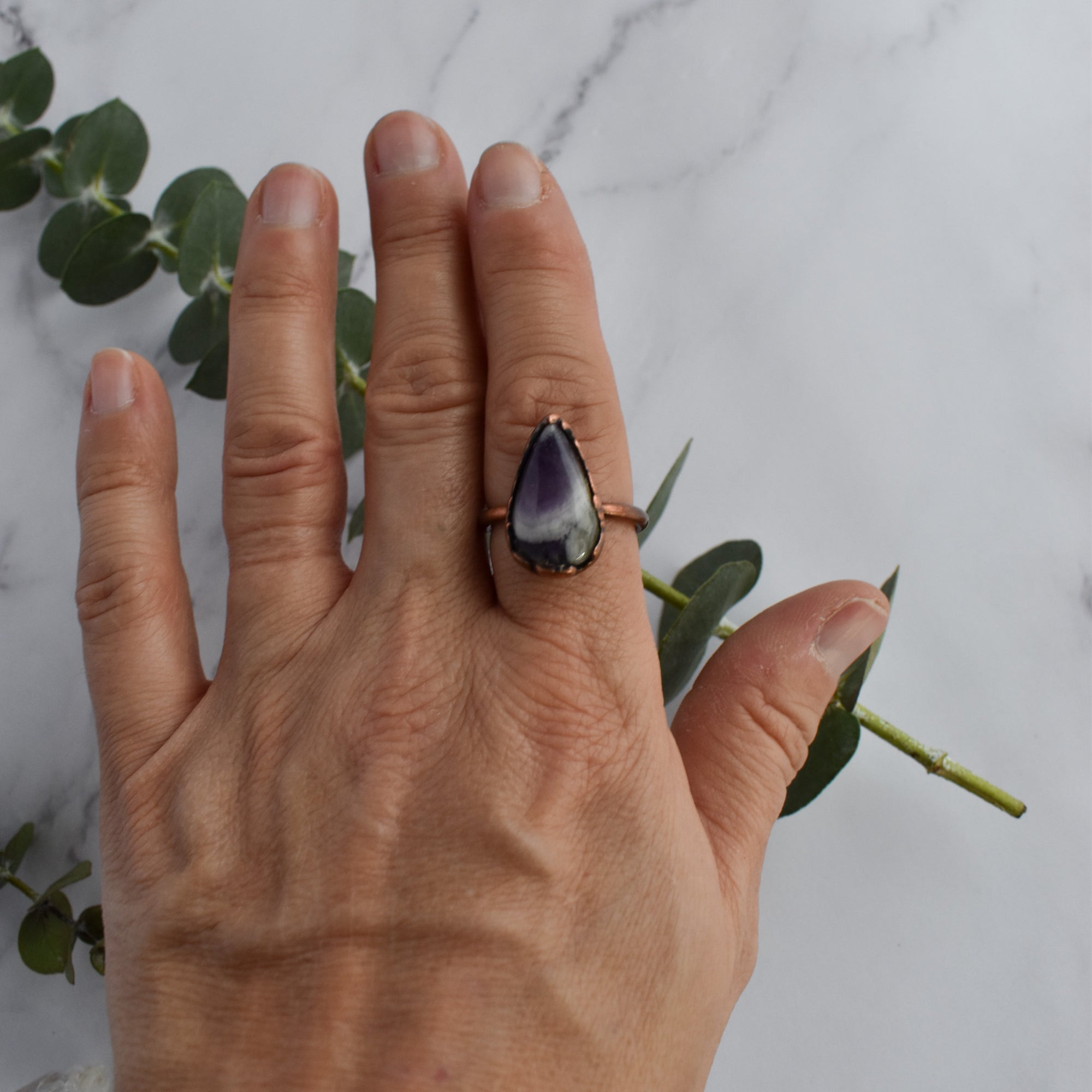 Tear drop shaped Chevron Amethyst ring worn on human hand. Green eucalyptus leaves in background.