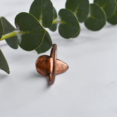 Light reflects off the shiny copper back of a ring. Green eucalyptus leaves are arranged in the background.