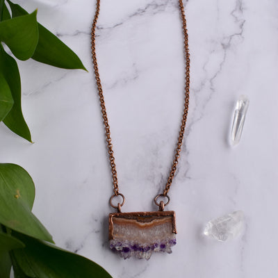 Shiny copper frames an Amethyst slice pendant. The banded layers show the crystal formation from brown and cream Agate to clear Quartz to sparkly purple and clear points. Pendant hangs from bright copper chain. Green leaves and Quartz points are arranged around necklace.