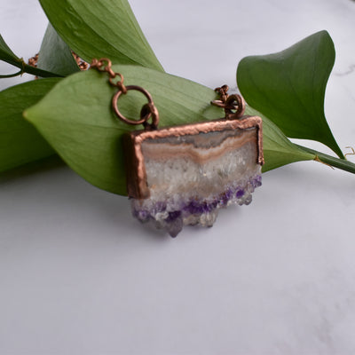 Shiny copper frames an Amethyst slice pendant. The banded layers show the crystal formation from brown and cream Agate to clear Quartz to sparkly purple and clear points. Pendant hangs from bright copper chain. Pendant is draped over green leaves.