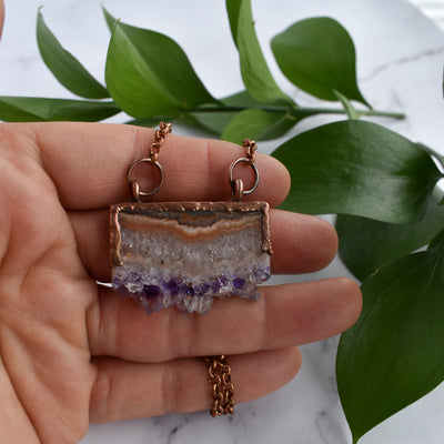 Shiny copper frames an Amethyst slice pendant. The banded layers show the crystal formation from brown and cream Agate to clear Quartz to sparkly purple and clear points. Pendant hangs from bright copper chain. Green leaves are arranged in background and a hand is holding the pendant.