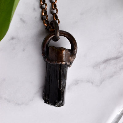 A Black cylindrical Tourmaline stone in an antiqued copper setting, the pendant hangs from a copper chain. Green leaves peaks from the corner.