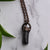 A Black cylindrical Tourmaline stone in an antiqued copper setting, the pendant hangs from a copper chain. Green leaves peak from the corners.