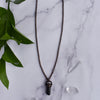 A Black cylindrical Tourmaline stone in an antiqued copper setting, the pendant hangs from a copper chain. Green leaves and Quartz points are arranged beside the necklace.