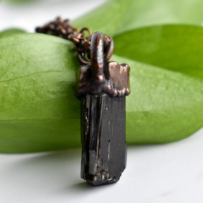 A Black cylindrical Tourmaline stone and antiqued copper pendant is arranged over green leaves.