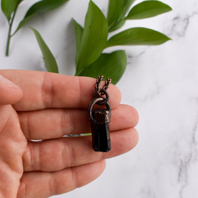 A Black cylindrical Tourmaline stone and antiqued copper necklace is held in a human hand. Green leaves are arranged in the background.