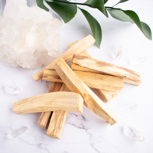 A stack of Palo Santo sticks are displayed next to quartz crystals and a green stem of leaves.