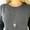 A woman in a black shirt wears a bluey green Labradorite coffin pendant. A rainbow moonstone accents the top of the copper bail, and hangs on a pure copper rolo chain.