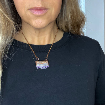 Shiny copper frames an Amethyst slice pendant. The banded layers show the crystal formation from brown and cream Agate to clear Quartz to sparkly purple and clear points. Pendant hangs from bright copper chain. Necklace is worn by woman in black shirt, the necklace length rests just below the collar bones.