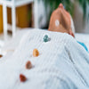 A woman lays on a table covered in a white blanket. Red, orange, yellow, green crystals are placed on top of her body at her Chakra points.